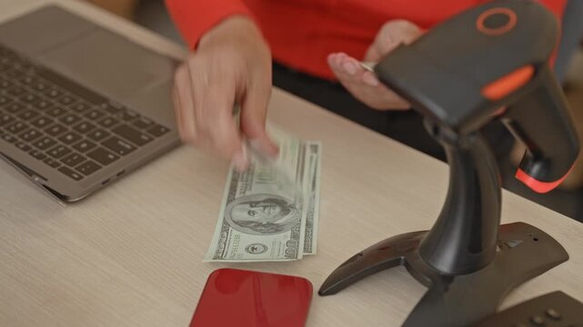 Woman counts cash with hands beside laptop and barcode scanner in building; confidence small business.