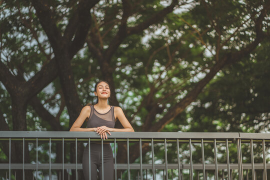 A young Asian woman in grey sportswear leaning on a park railing with eyes closed, enjoying a peaceful rest after exercise. Concept of wellness, mindfulness, and healthy lifestyle during a workout bre