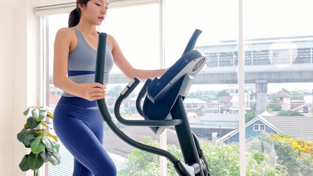 Woman exercising on an elliptical trainer at home beside large windows, wearing athletic wear, with urban skyline and rail station outside.