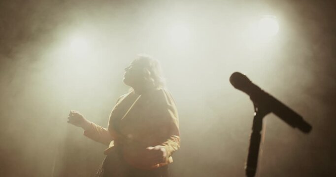 A female singer dancer performs on a dark stage with bright backlights, haze, and a microphone stand, creating a dramatic concert atmosphere