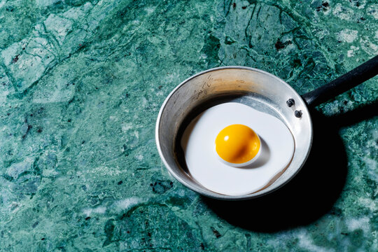 Surreal glossy fried egg illusion in toy metal pan on green marble