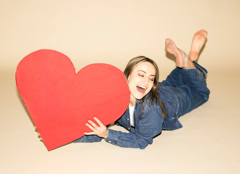Woman holding a red paper heart while lying down smiling in jeans outfit