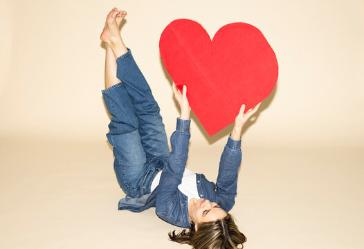Woman holding a red paper heart while lying down in a jeans outfit