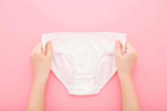 Little child girl hands holding and showing white panties on light pink table background. Pastel color. Closeup. Point of view shot. Top down view.