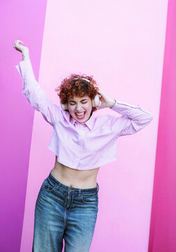Cheerful redhead woman dancing to music with headphone by pink wall