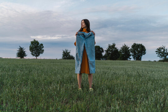 Woman holding blue denim pants standing in corn field at sunset