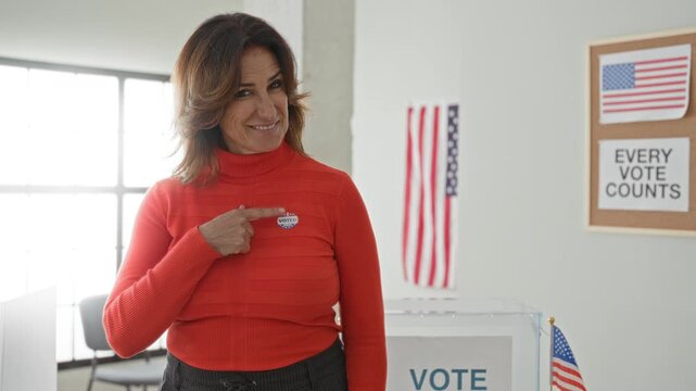 Woman points finger to voted sticker on chest near transparent ballot box and american flag in polling building; pride civic duty.