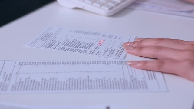 Focused businesswoman hand holding pen near detailed financial document and invoice paper report during serious stressful corporate accounting analysis reviewing printed data carefully on white desk