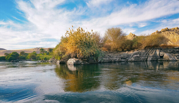 Panoramic nature landscape of the River Nile featuring lush green trees and bushes on the rocky banks reflected in calm water. A bright spring and summer travel scene in Egypt.