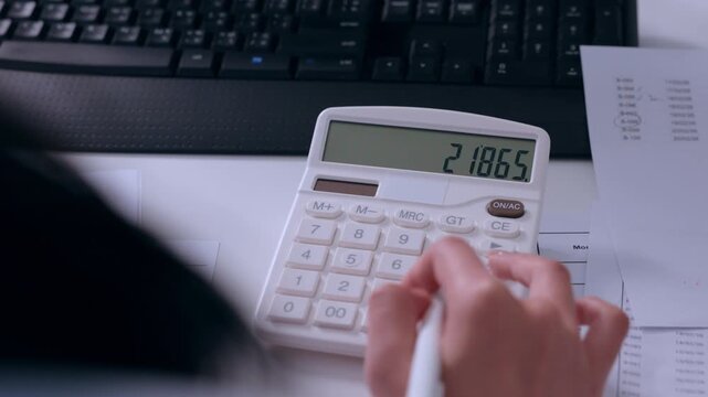 Focused person calculating business finance paperwork using white calculator near computer keyboard examining tax document and accounting report on modern office desk showing concentration
