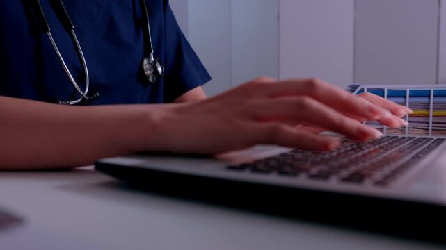 Medical professional in blue scrub maintaining deep focus to type online patient data on laptop keyboard while working late inside dark hospital clinic with stethoscope around neck for healthcare