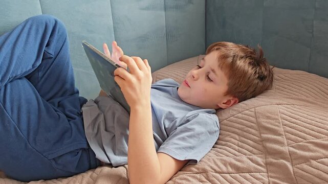 Boy lying on back holding tablet up. comfortable position, reading or gaming, home relaxation, digital device.