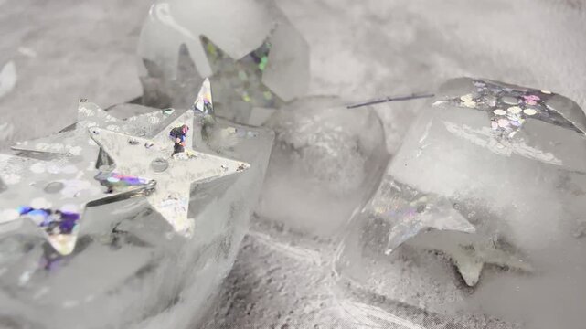 Macro video of glittery silver star sequins on melting ice with pouring water and dreamy reflections for luxury, festive, or beauty visuals.