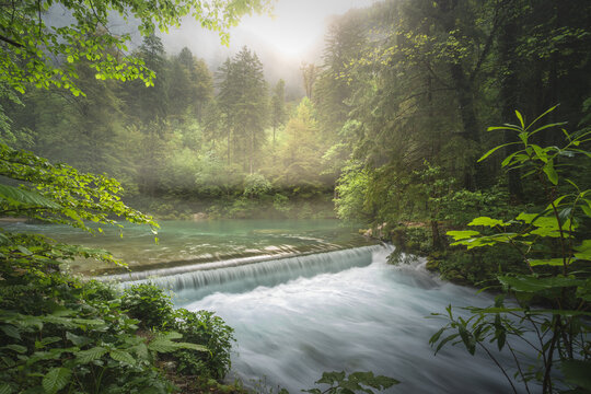 View of a serene river cascading over a small weir, its emerald waters flowing through a vibrant, misty forest bathed in ethereal sunlight, Stahovica, Kamnik, Slovenia.