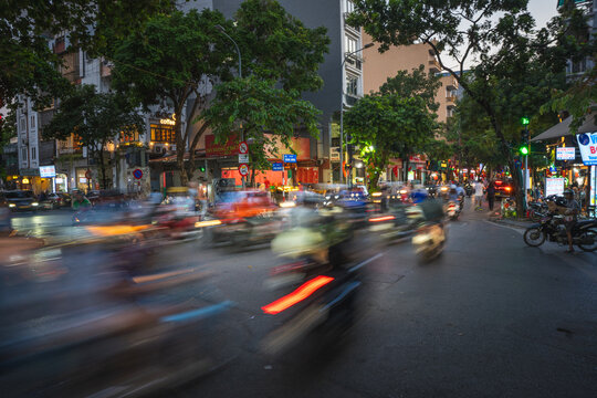 Ha Noi, Vietnam - 07 October 2024: View of a bustling street at dusk, where vibrant streaks of red and white vehicle lights blur through the dark, tree-lined urban landscape.