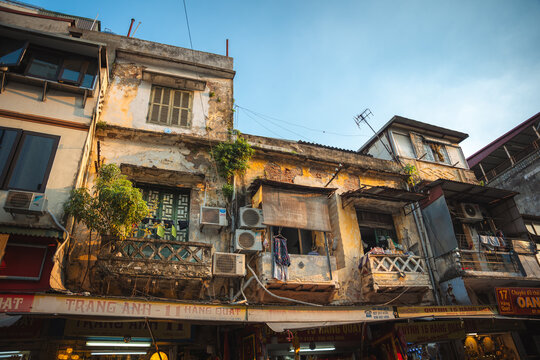 Ha Noi, Vietnam - 07 October 2024: View of weathered colonial-era buildings with peeling facades, intricate balconies, and bustling street-level shops under a clear sky.