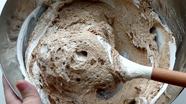 A person kneads the halva ingredients in a bowl with a spatula, preparing it. Various ingredients are visible in the finished mixture. Sunflower halva.