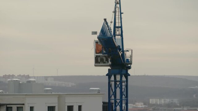 Industrial tower crane operating on a construction site, with its long blue jib slowly rotating against a grey, overcast sky overlooking a sprawling city landscape in the background