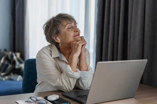 Dreamy senior woman sitting at table with laptop listening to music or podcast in headphones.