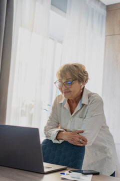 Happy senior woman in headphones standing by chair looking at laptop screen at home