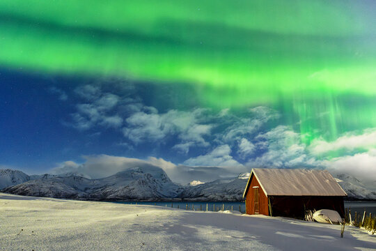 View of a vibrant aurora borealis painting the sky above a rustic red barn nestled in a snowy landscape against a backdrop of majestic mountains, Tromso, Troms, Norway.