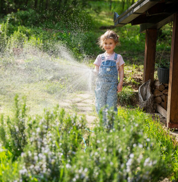 Happy little girl in denim overalls watering green lawn with garden hose on a sunny summer day