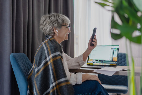 Happy senior woman in headphones having video call on smartphone wrapped in blanket.