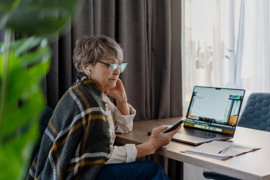 Cozy senior woman wrapped in blanket sitting at table and looking at smartphone screen.