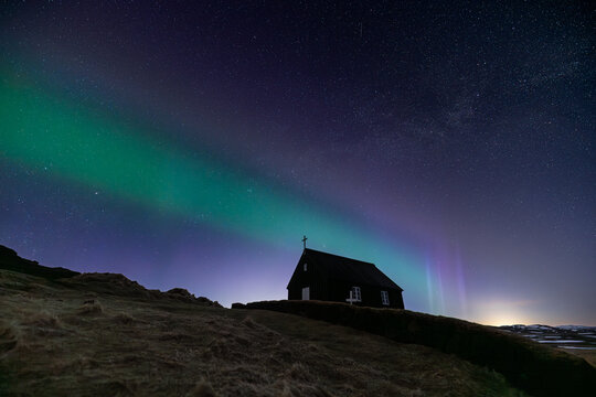 View of a small church silhouette against the vibrant green and purple aurora borealis dancing in the star-filled night sky, Krysuvikurkirkja, Iceland.