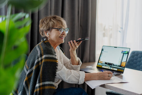 Social senior woman wrapped in blanket recording voice message on smartphone at home
