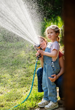 Happy mother and little daughter in denim overalls laughing and watering lawn with hose together.
