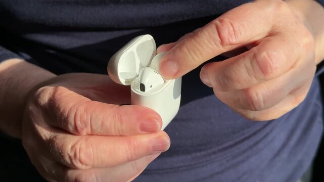 Closeup woman holding white earbud case, gently removing single earbud for listening warm sunlight highlights textured hands