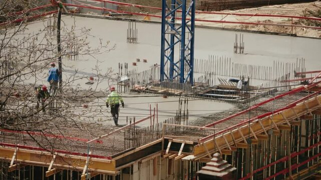 High-angle view of construction workers pouring and leveling a wet concrete slab for a new building foundation, working around reinforcing steel bars on an active building site. timelapse