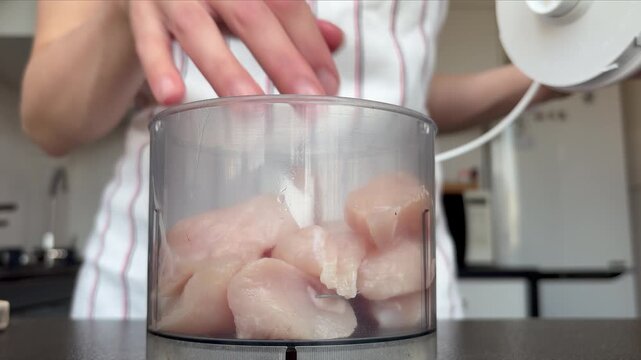 A housewife in an apron uses a blender to chop chicken fillet at home in the kitchen