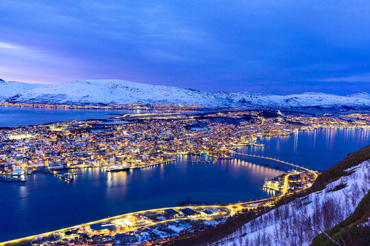 View of sparkling city lights illuminating a serene deep blue fjord, framed by majestic snow-dusted mountains under a twilight glow, Tromsdalen, Troms, Norway.