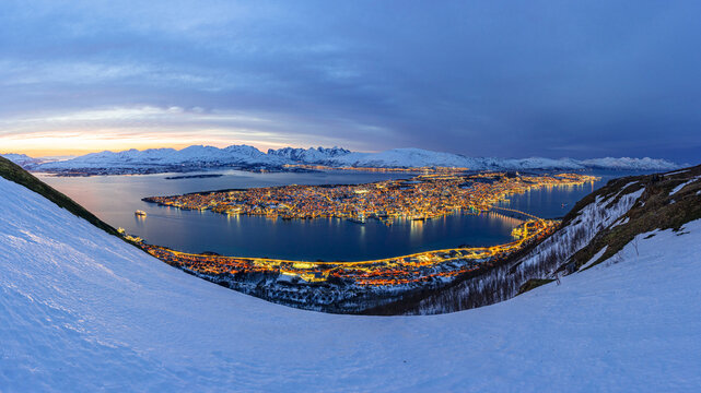 View of a vibrant island city sparkling with golden lights, contrasting with the serene blue twilight sky and pristine snow-covered slopes, Tromsdalen, Troms, Norway.