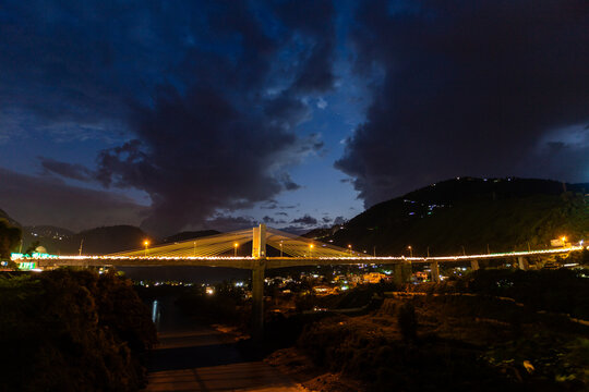 View of a bridge illuminated at night, cutting through the darkness with streaks of light, against a backdrop of mountains and a twilight sky, Muzaffarabad, Pakistan.