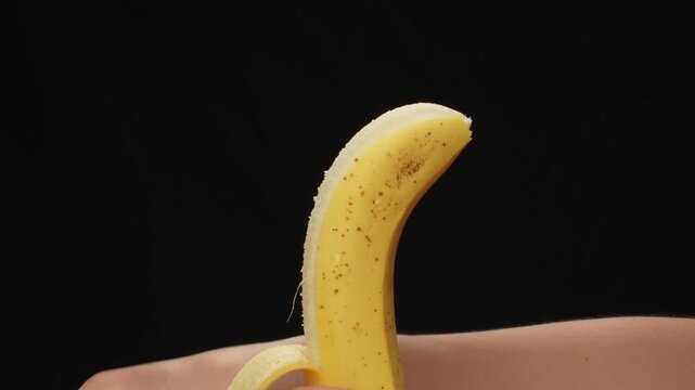 Close-up of a woman's hand peeling a bright ripe yellow banana, standing out against a contrasting black background.