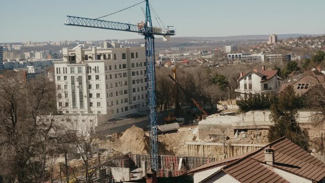 Blue tower crane rotating its jib over an urban construction site with a new building and foundation pit, showing the process of urban development and real estate investment on a sunny day. timelapse