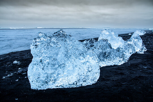 View of shimmering ice diamonds scattered on the stark black sands, a cold, surreal landscape under a muted sky, Glacier Lagoon, Sveitarfelagid Hornafjordur, Iceland.