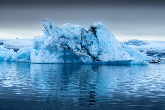 View of a luminous blue iceberg drifts serenely in the tranquil lagoon, reflecting the muted sky, a dance of light and shadow, Glacier Lagoon, Iceland.
