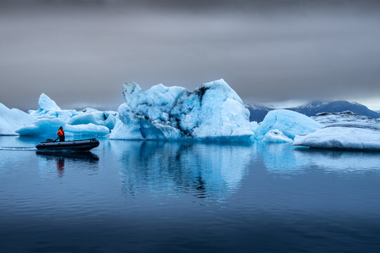 View of a lone boat navigates through the serene, icy waters amidst towering icebergs under a brooding sky, Glacier Lagoon, Sveitarfelagid Hornafjordur, Iceland.