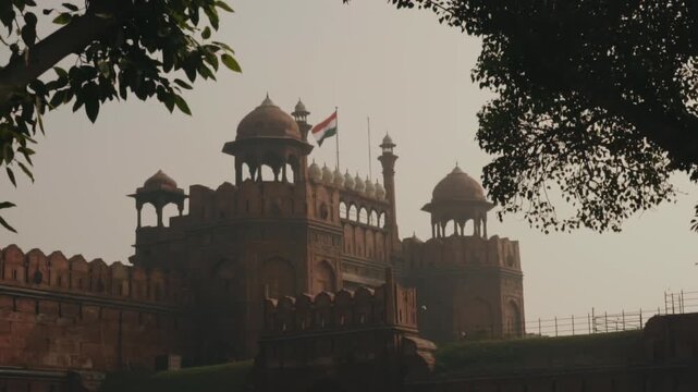 Majestic view of the historic Red Fort, a UNESCO World Heritage Site in Delhi, with the Indian national flag waving proudly against a hazy sky, framed by tree leaves in the foreground