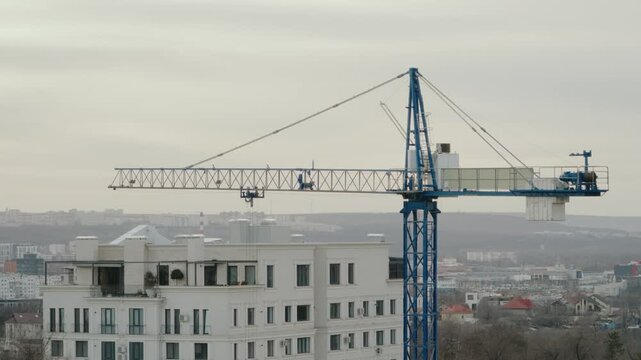 Industrial tower crane operating on a construction site, with its trolley moving along the horizontal jib against a backdrop of the city skyline and an apartment building on an overcast day