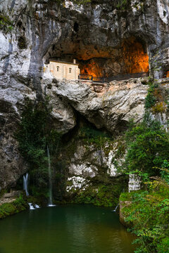 View of a glowing chapel in a cave, above a green pool and waterfall, Santa Cueva de Covadonga, Cangas de Onis, Spain.