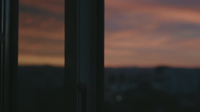 Anonymous man's silhouette closing a window handle at home, with a beautiful and colorful urban sunset visible outside. A contemplative and moody scene reflecting on the end of the day