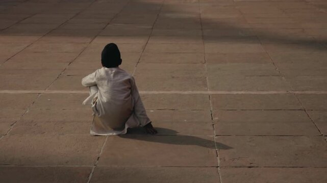 Lonely Indian child wearing traditional clothing sitting barefoot on the sunlit stone floor of a temple courtyard, casting a long shadow in a contemplative and peaceful moment of solitude