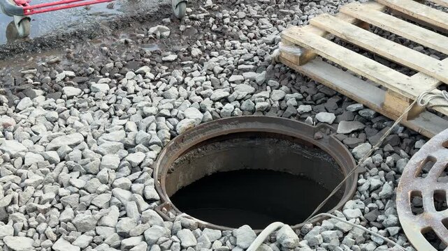 Open inspection well with rubber hose, gravel street, wooden pallet and shopping cart, pump engine running as hose drains water