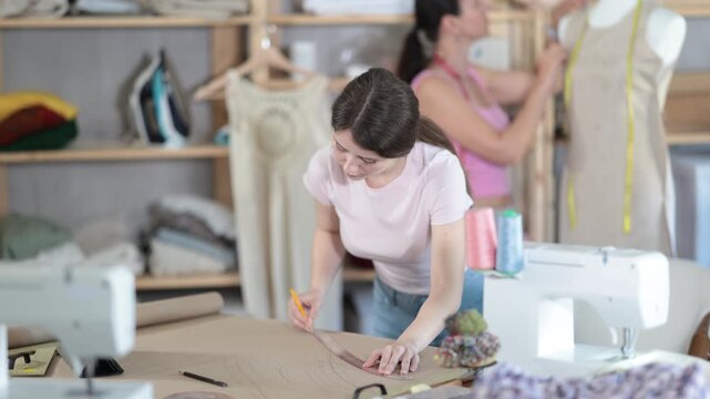 Woman tailor draws dress diagram scheme on paper. Companion worker in background with mannequin. High quality 4k footage