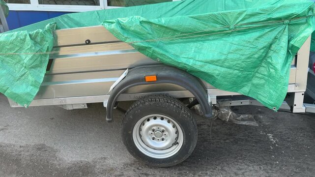 Small car trailer covered green tarp, parked roadside on asphalt near dealership, visible wheel, fender and hitch, light rust
